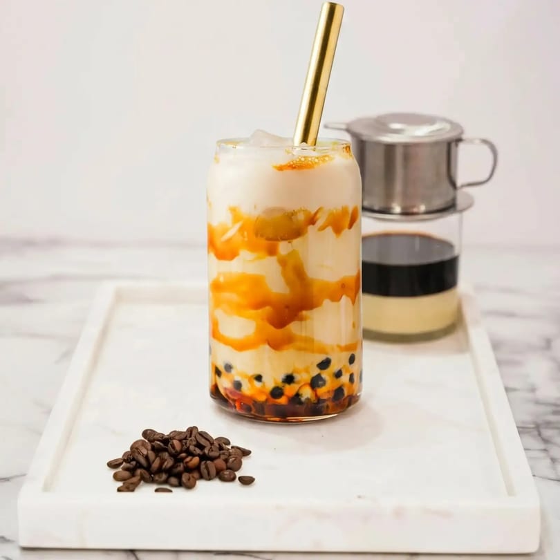 Boba tea with caramel swirls and a gold straw, next to a Vietnamese coffee filter and beans.