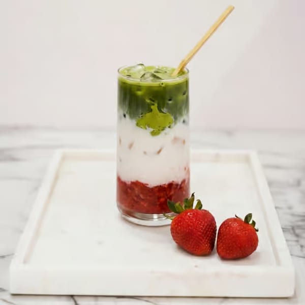 Layered strawberry, milk, and matcha drink with a straw and two strawberries on a marble tray.