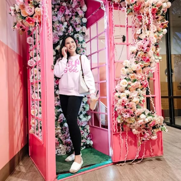 A smiling woman in a pink Barbie sweatshirt stands in a pink phone booth adorned with flowers.