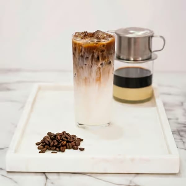 Iced Vietnamese coffee, a phin filter, and coffee beans on a white marble tray.