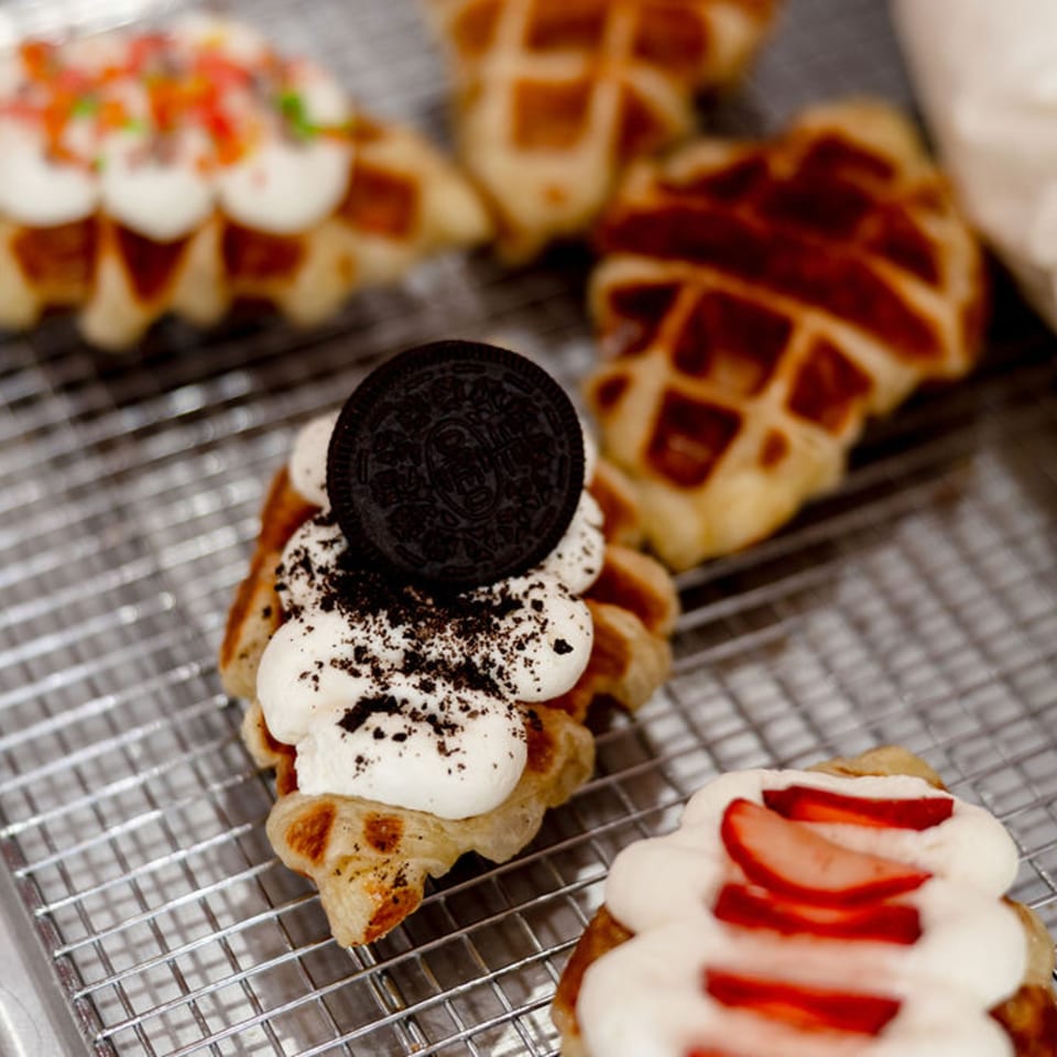 Croffles with various toppings, including whipped cream, Oreo, sprinkles, and strawberries, on a cooling rack.