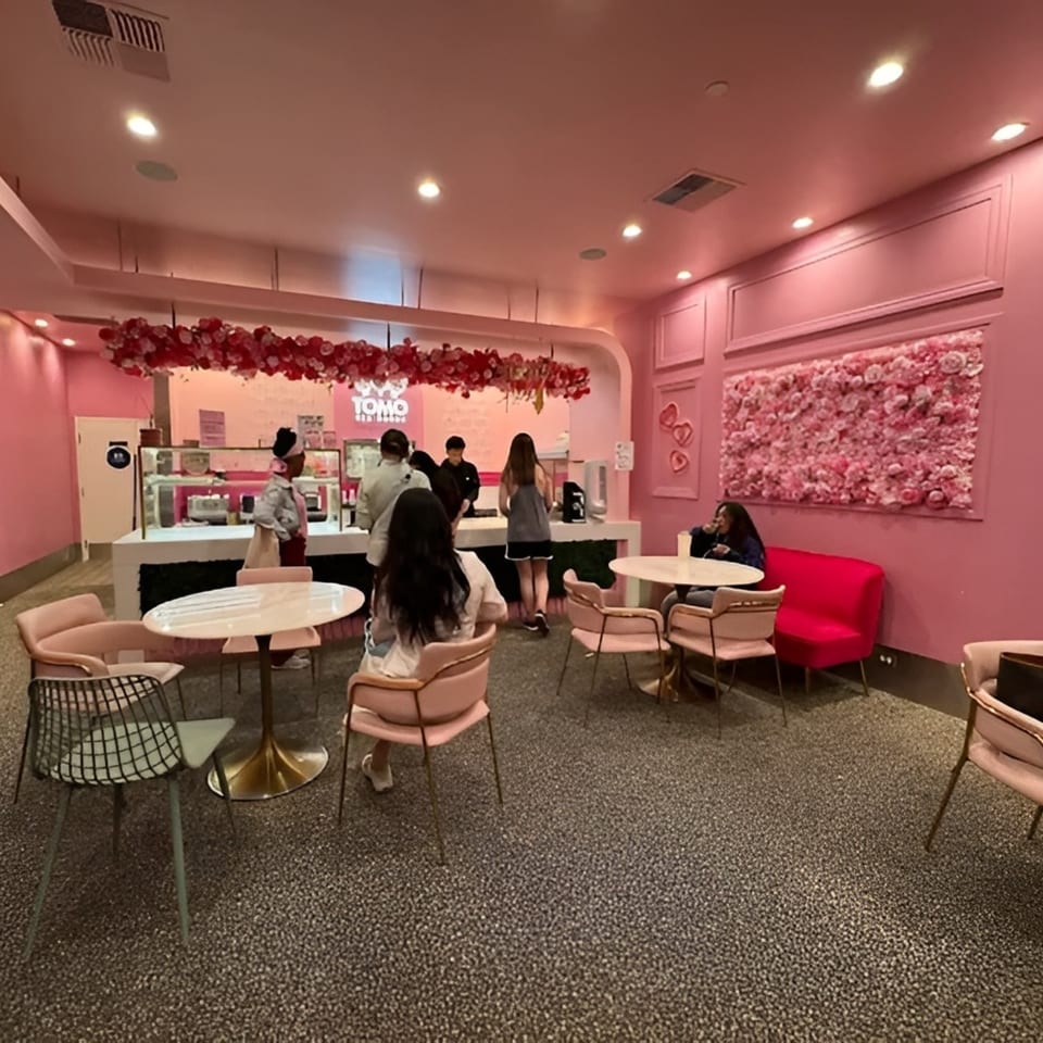 Pink-themed dessert shop with customers at the counter and seated at tables, adorned with floral decorations.