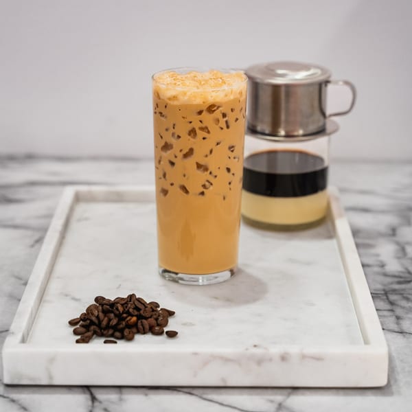Iced coffee with a Vietnamese phin filter, condensed milk, and coffee beans on a marble tray.